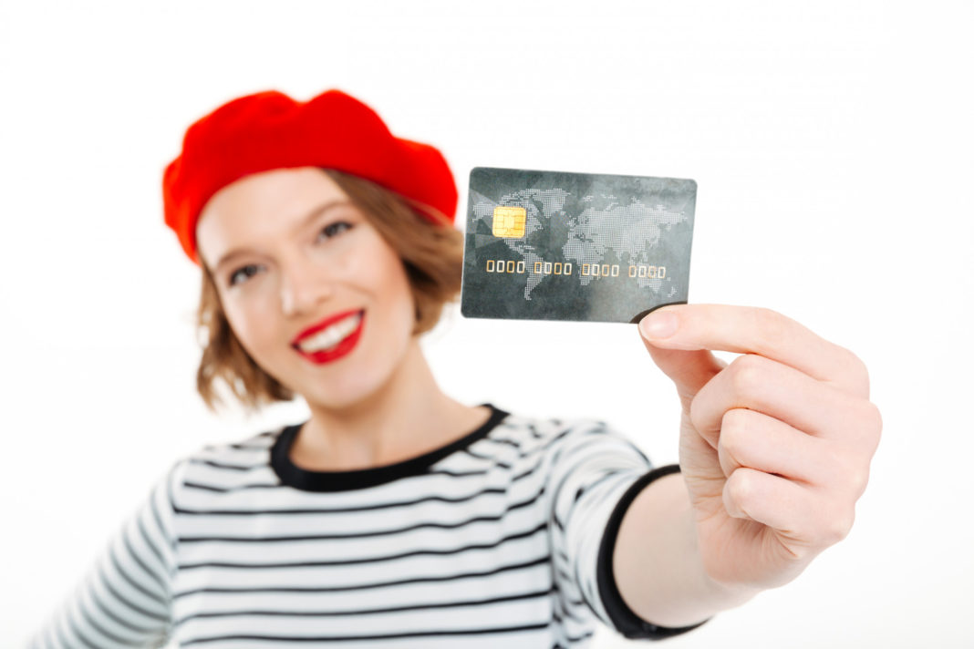 smiling-ginger-woman-showing-credit-card-camera-grey-focus-card